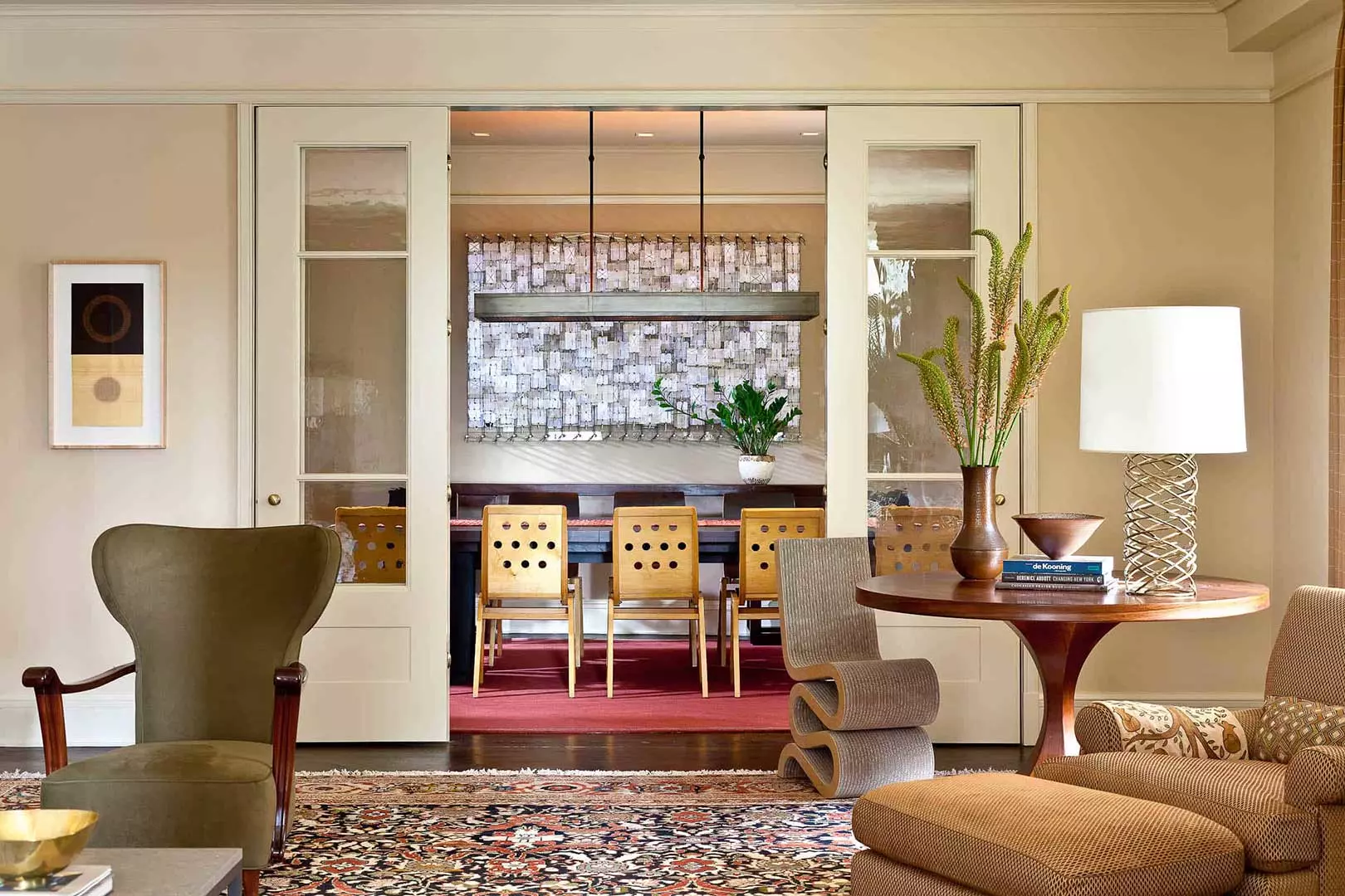 An eclectic array of furniture marks the transition from living room to dining room, the latter seen through wide French doors. In the Living Room, an antique Italian armchair by Ulrich Guglielmo, Italy from the 1950's echoes the curves of the Danish Modern rosewood pedestal table, the Frank Gehry Wiggle Chair, and Herve van der Straeten's "Tornade" lamp.
