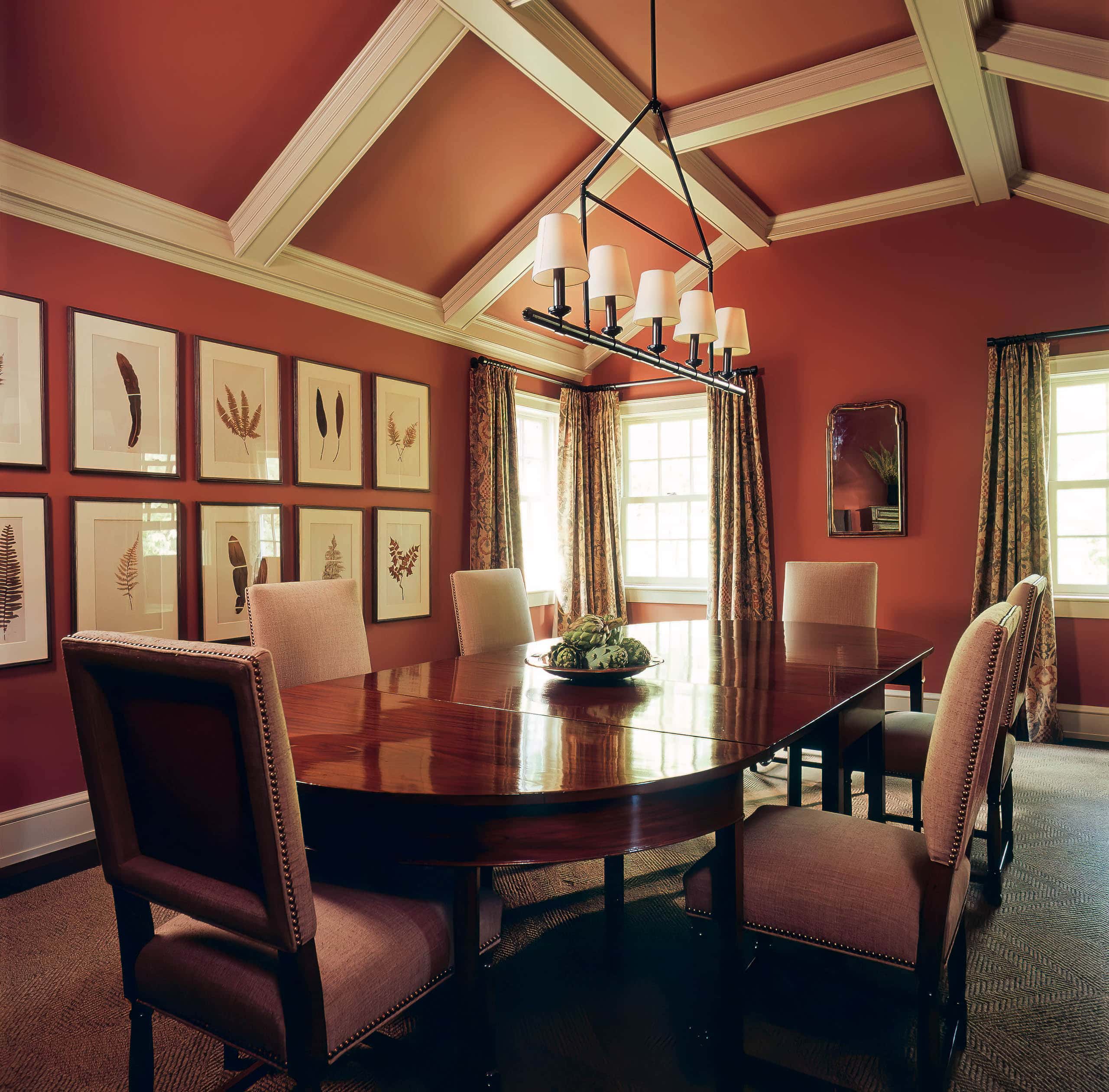 The walls and inset panels of the arched and coffered ceiling in this dining room were painted in Farrow & Ball's Loggia No. 232, setting the tone for lively conversation. In contrast to the rich russet-red, a series of framed botanicals reference the gardens on the property beyond the glass.